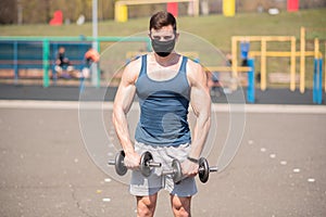 Young sports guy doing exercises with dumbbells in a medical mask on the playground during a pandemic