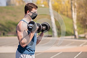 Young sports guy doing exercises with dumbbells in a medical mask on the playground during a pandemic