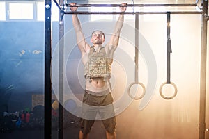 Young sports man with heavy waistcoat makes pull ups on crossbar in the gym full of smoke.