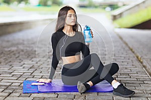 A young sports woman leading a healthy lifestyle, rests after training, drinks water and listens to music