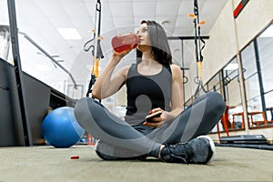 Young sports woman resting on the floor after exercises in gym, drinking water reading smartphone. Fitness, sport, training,
