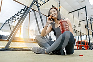 Young sports woman resting on the floor after exercises in gym, drinking water talking on smartphone. Fitness, sport, training,