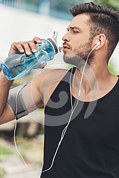 young sportsman in earphones with smartphone in running armband case drinking