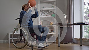 Young sportsman in wheelchair throwing basketball ball in slow motion training at home indoors. Portrait of inspired