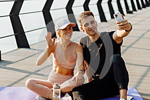 Young sportsmen man and woman resting after workout taking selfie on mobile phone sitting on fitness mat outdoors