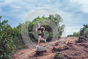 Young sportswoman cross country running on dirty rocky footpath in mountains in summer. Fit girl jogging outdoors in