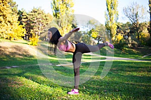 Young sportswoman doing warm-up balance exercise standing on one legs stretching leg and back outdoors. Warm-up stretching at the