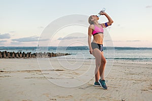 young sportswoman in earphones with smartphone in armband case drinking water from bottle on beach with sea