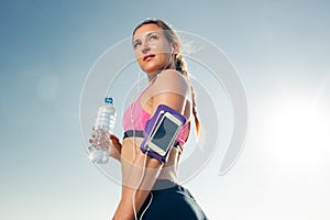 young sportswoman in earphones with smartphone in armband sport case holding bottle of water against blue