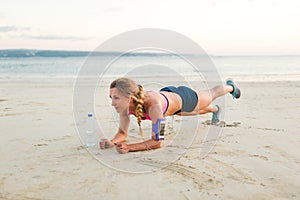 young sportswoman in earphones with smartphone in running armband case and bottle of water doing plank on sandy