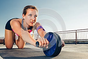 young sportswoman in earphones with smartphone in running armband case doing exercise with abs roller on fitness mat on rooftop