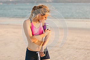 young sportswoman in earphones using smartphone in running armband case on beach with sea