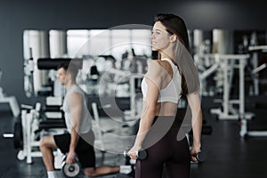 Young sportswoman lifting weights in gym wearing sportswear with her boyfriend on background