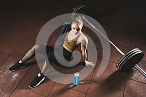 Young sportswoman with towel on shoulder sitting on floor near barbell