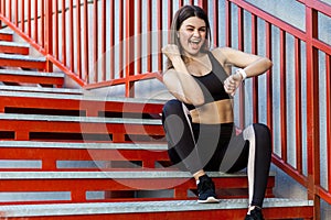 Young sporty beautiful woman having a break sitting on the red stairs after morning jogging, checking on smartwatch and celebrate