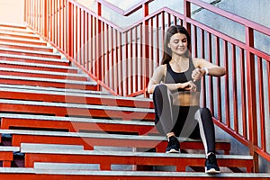 Young sporty beautiful woman having a break sitting on the red stairs after the morning jogging in the modern city, checking
