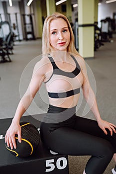 Young sporty blond woman in black and pink top and black leggings sitting on exercise bench and holding ball in gym.