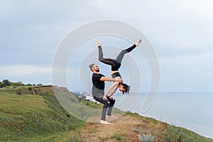 Young sporty couple wearing black pants, t-shirts and topic practicing acroyoga handstand at sunrise or sunset on the edge of the