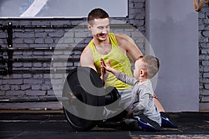 Young sporty father and little cute son sitting near barbell against brick wall in the cross fit gym.