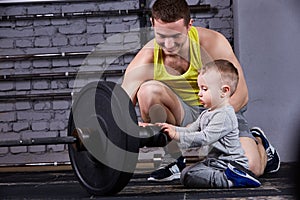 Young sporty father showing dumbbells for his little son and smiling against brick wall in the cross fit gym.