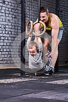 Young sporty father trains the little son with gymnastic rings against brick wall at the cross fit gym.