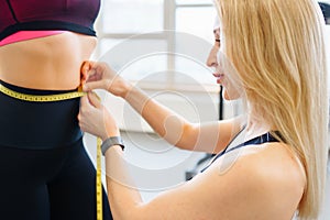 Young sporty female personal trainer in black top measuring woman& x27;s waist to track progress in gym. Fitness concept