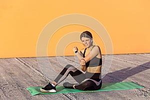 Young sporty girl in black sportwear sitting on mat and have a strong hurt problem with elbow, spasm painful. Gripping cubit with