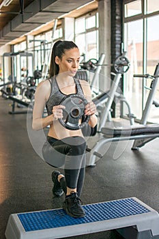 Young sporty girl doing exercise with weight barbell plate on fitness stepper in gym