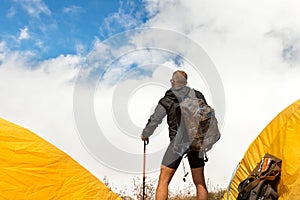 Young sporty guy with a backpack in a tent camp looking afar against a background of cumulus clouds and a blue sky.