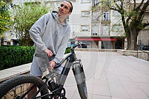 Young sporty man adjusts the height of new battery powered bike, electric bicycle before taking it out for first ride