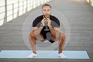 Young sporty man in black fitwear squatting holding dumbbell outdoor