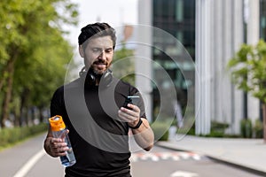 A young sporty man with headphones is standing on a city street, holding a bottle of water and using a mobile phone