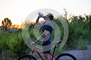 Young sporty man riding a bicycle drinking water from the sport bottle at sunset