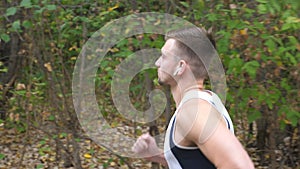 Young sporty man running along trail near forest at early autumn. Handsome sportsman with wireless headphones jogging