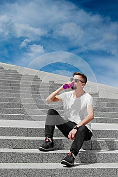 Young sporty man sitting on stairs outdoors drinking water from eco plastic bottle against blue sky background.