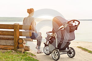 Young sporty mother with stroller sitting on wooden bench near lake or river. Mom walking with baby in pram near pond at early mor