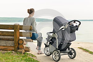 Young sporty mother with stroller sitting on wooden bench near lake or river. Mom walking with baby in pram near pond at early mor