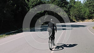 Young sporty woman is biking on forest road in sunny day. Professional cyclist in black cycling outfit is cycling on empty car roa