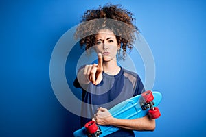 Young sporty woman with curly hair and piercing holding skate over blue background pointing with finger to the camera and to you,