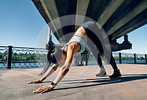 Young sporty woman doing yoga asana downward facing dog under industrial bridge