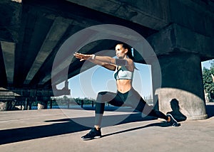 Young sporty woman doing yoga asana Warrior I Pose outdoors under industrial bridge