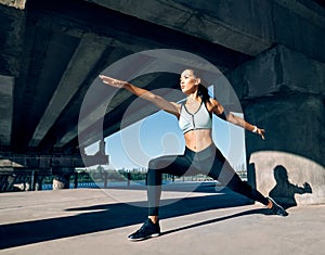 Young sporty woman doing yoga asana Warrior I Pose outdoors under industrial bridge