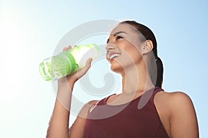 Young sporty woman drinking water from bottle against blue sky