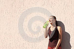 Young sporty woman drinking water from bottle near wall outdoors on sunny day