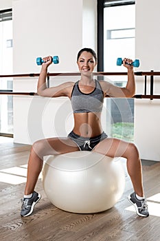 Young sporty woman in gym doing fitness exercise with white ball and dumbbells