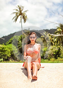 Young sporty woman in orange bikini and sunglasses sits on fine beach sand, wind in her hair, palm tree and jungle behind her