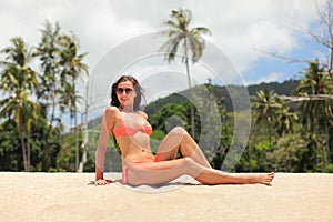 Young sporty woman in orange bikini and sunglasses, sitting on the sand near beach, palm trees behind her