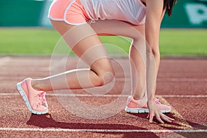 Young sporty woman in pink shorts and tank tops are ready to run and stands on start on stadium track. Close-up legs and arms