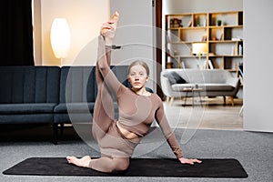 Young sporty woman practicing yoga, doing Compass pose, wearing sportswear pants and top sitting on the floor in living room