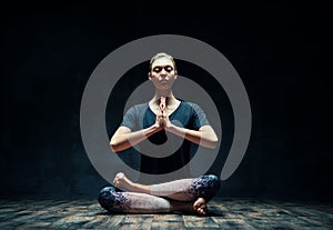 Young sporty woman practicing yoga sitting in lotus pose with namaste in dark room